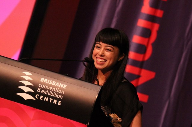 Slightly off-center (tilted to the right) image of a woman with dark hair smiling widely standing behind a podium that says Brisbane convention & exhibition centre.