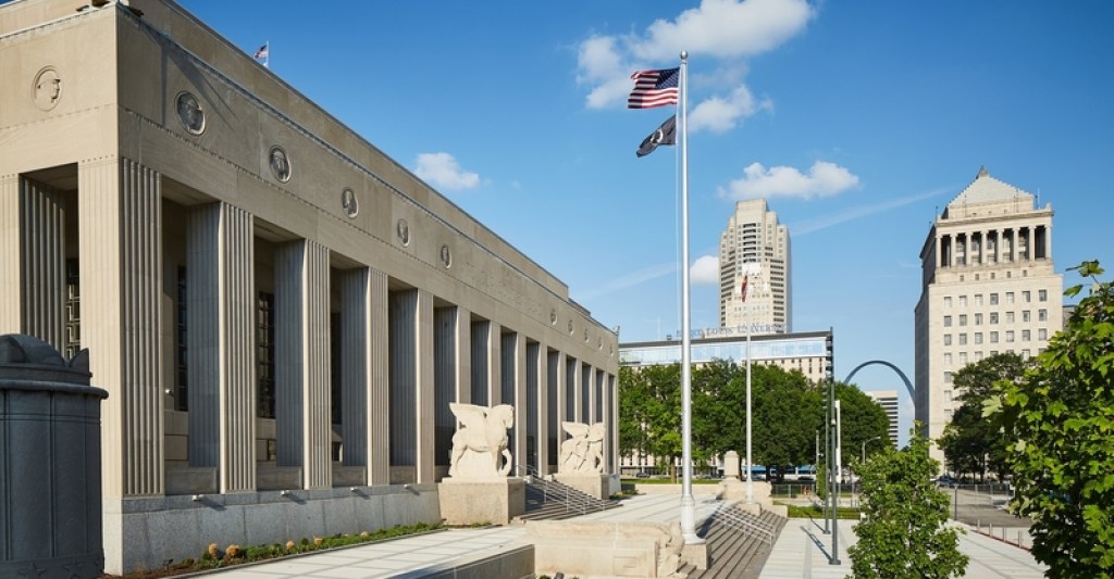 The facade of the Soldiers Memorial, a neoclassical structure with columns along the front and winged statues along the steps to the entrance. Other buildings and structures are visible in the background, including the famous Arch.