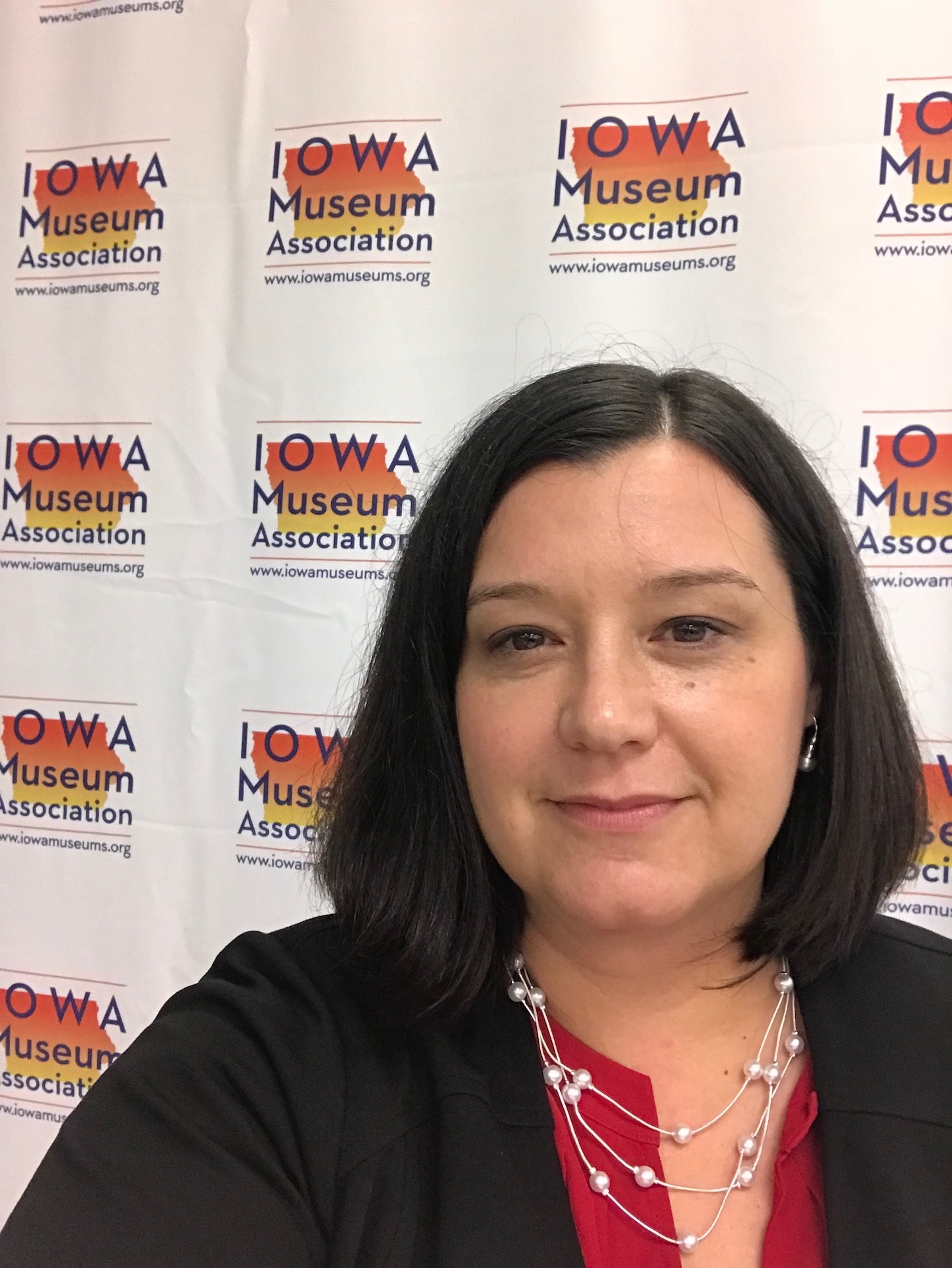 Image of a white woman standing in front of an Iowa Museums Association step and repeat. She has shoulder length brown hair combed to the left side wearing a dark colored jacket and red shirt.