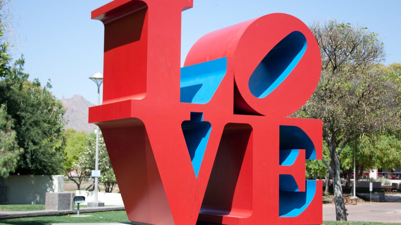 A sculpture of the famous "LOVE" logo by Robert Indiana, where the L and O are stacked on top of the V and E, and the O is slanted sideways. The sculpture is displayed outside with trees and a street visible in the background.