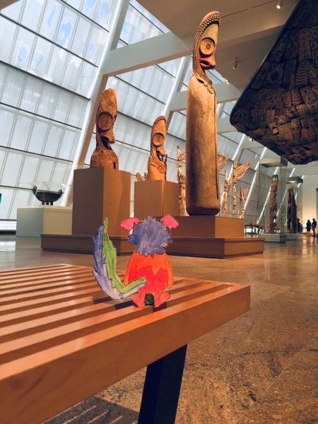 Marabou is seen from behind, perched on a slatted museum bench in front of a collection of wood scupltures from Oceania.