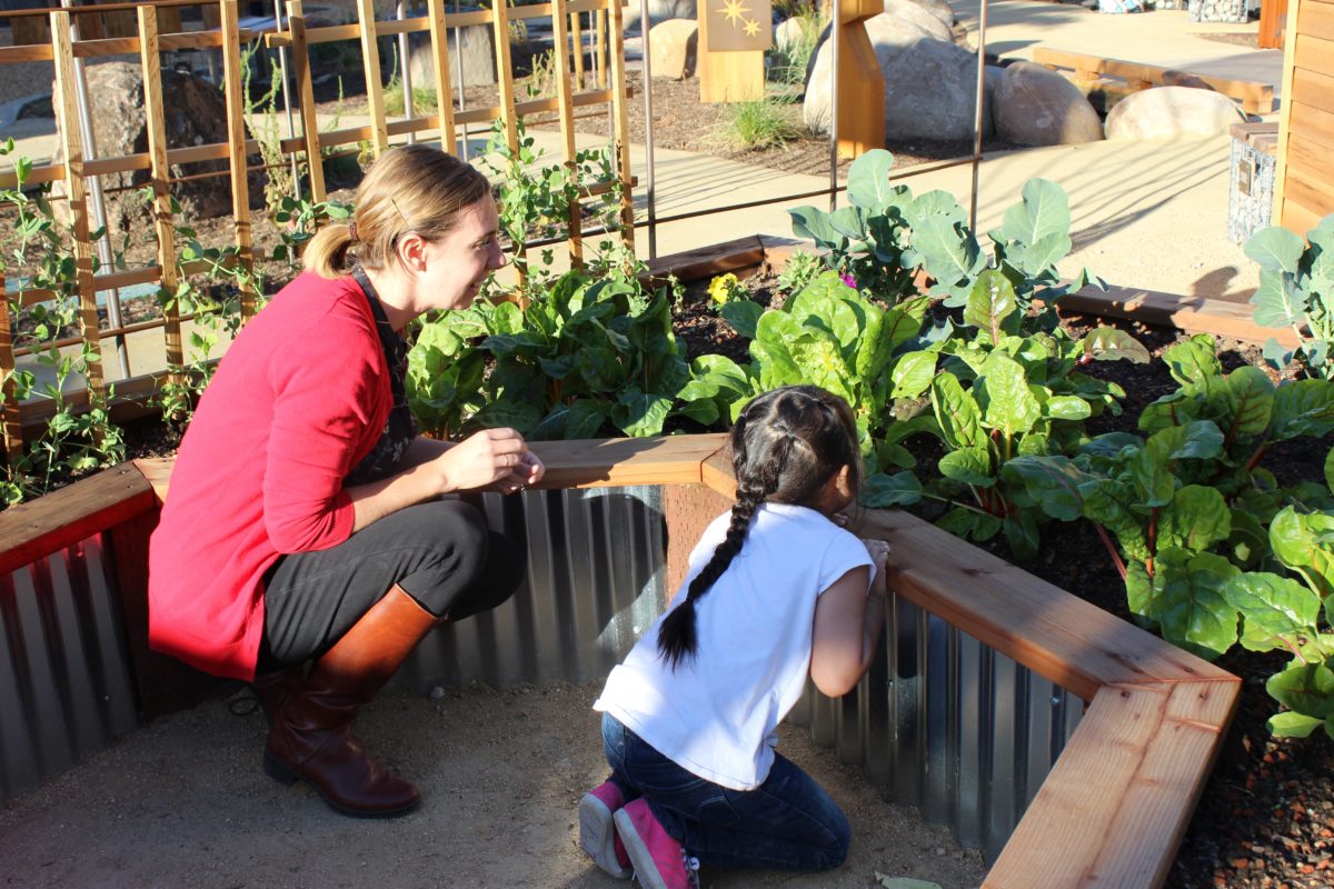 An adult woman crouches with a young visitor to look at vegetables growing in a garden.