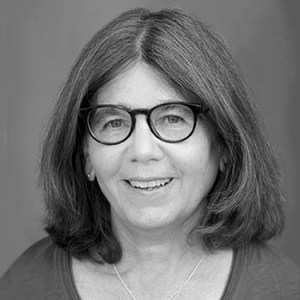 Black and white headshot of Joan Baldwin a white woman with shoulder length brown hair wearing dark rimmed glasses