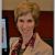 Headshot of Anne Basham standing in front of a computer which is off to the left hand side of the screen. She is a white woman with short cropped hair wearing a tan suit jacket and colorful patterned top.