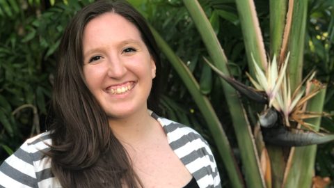 A portrait of a young woman smiling in front of a tropical plant.