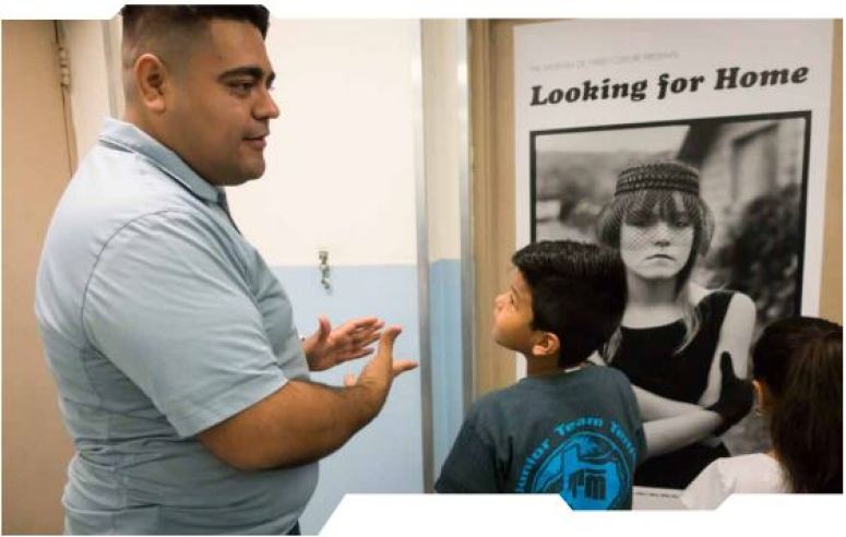 An Hispanic man talks to a young boy while looking at an image of a woman experiencing homelessness. 