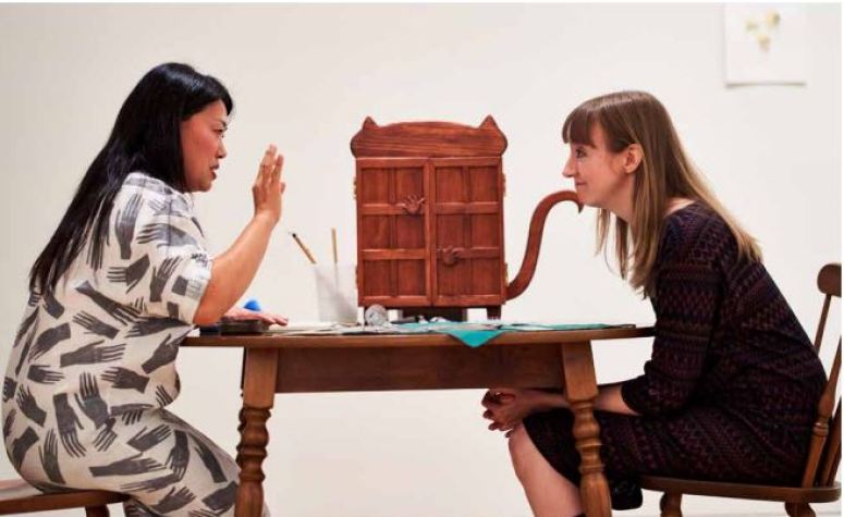Two women sits at a wooden table with a wooden case shaped like a cat sitting between them. 