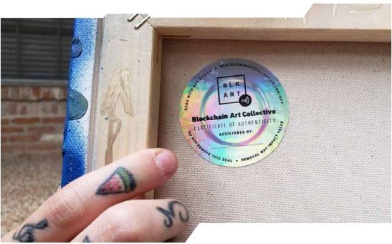A tattooed hand holds a painting by the stretcher, looking at the back there is an iridescent sticker for the Blockchain Art Collective. 