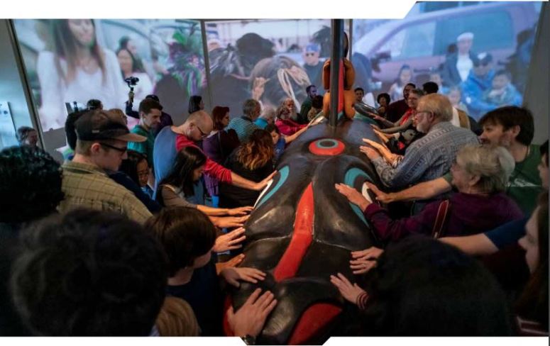 A large group of people stand with their hands on a whale totum in the exhibition. 