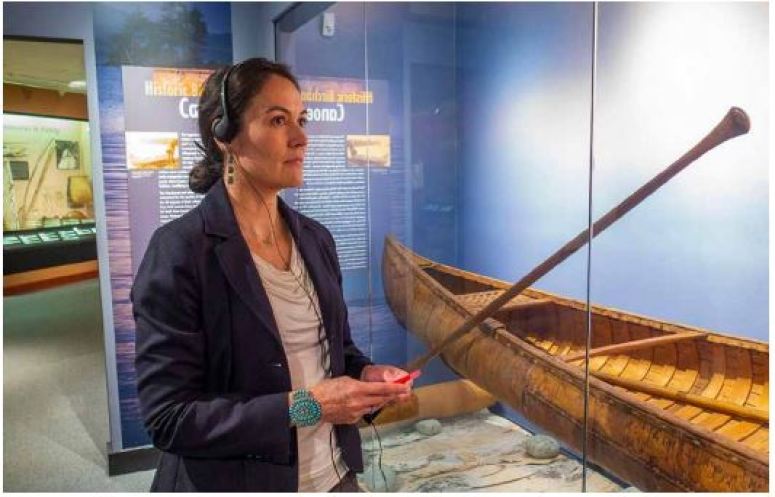 Shelly Lowe stands in front of a glass enclosed canoe on display with a set of headphones on. 