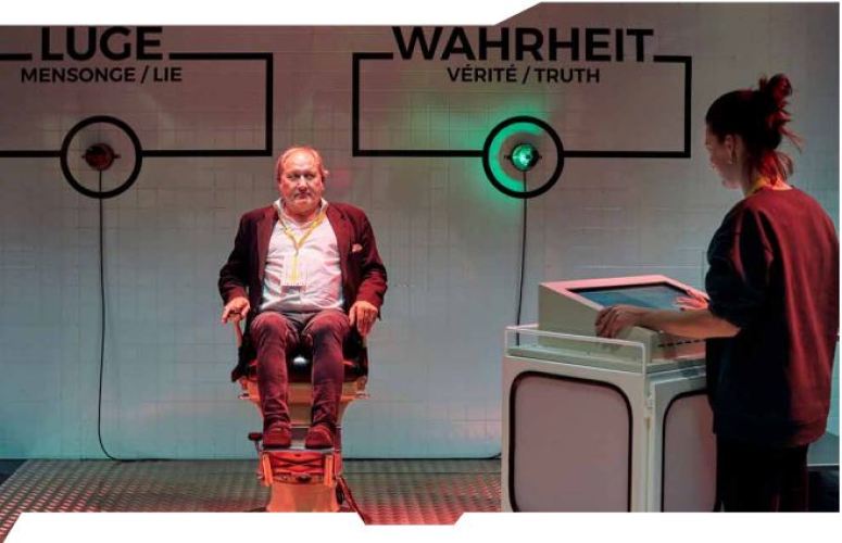 A gentleman sits on an elevated chair in an exhibit with a person standing off camera to the right at a podium with a green light shining in the background.