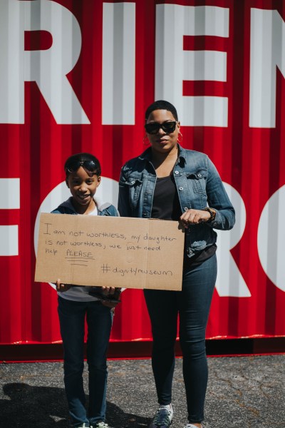 A woman and a child stand in front of the museum holding a cardboard sign that says "I am not worthless, my daughter is not worthless, we just need help PLEASE. #DignityMuseum"