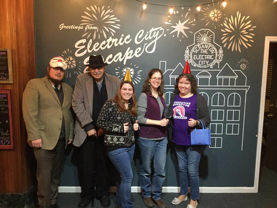 The staff of Luzerne County Historical Society pose for a group portrait in front of a wall mural that says "Greetings from Electric City Escape; Scranton, the Electric City."