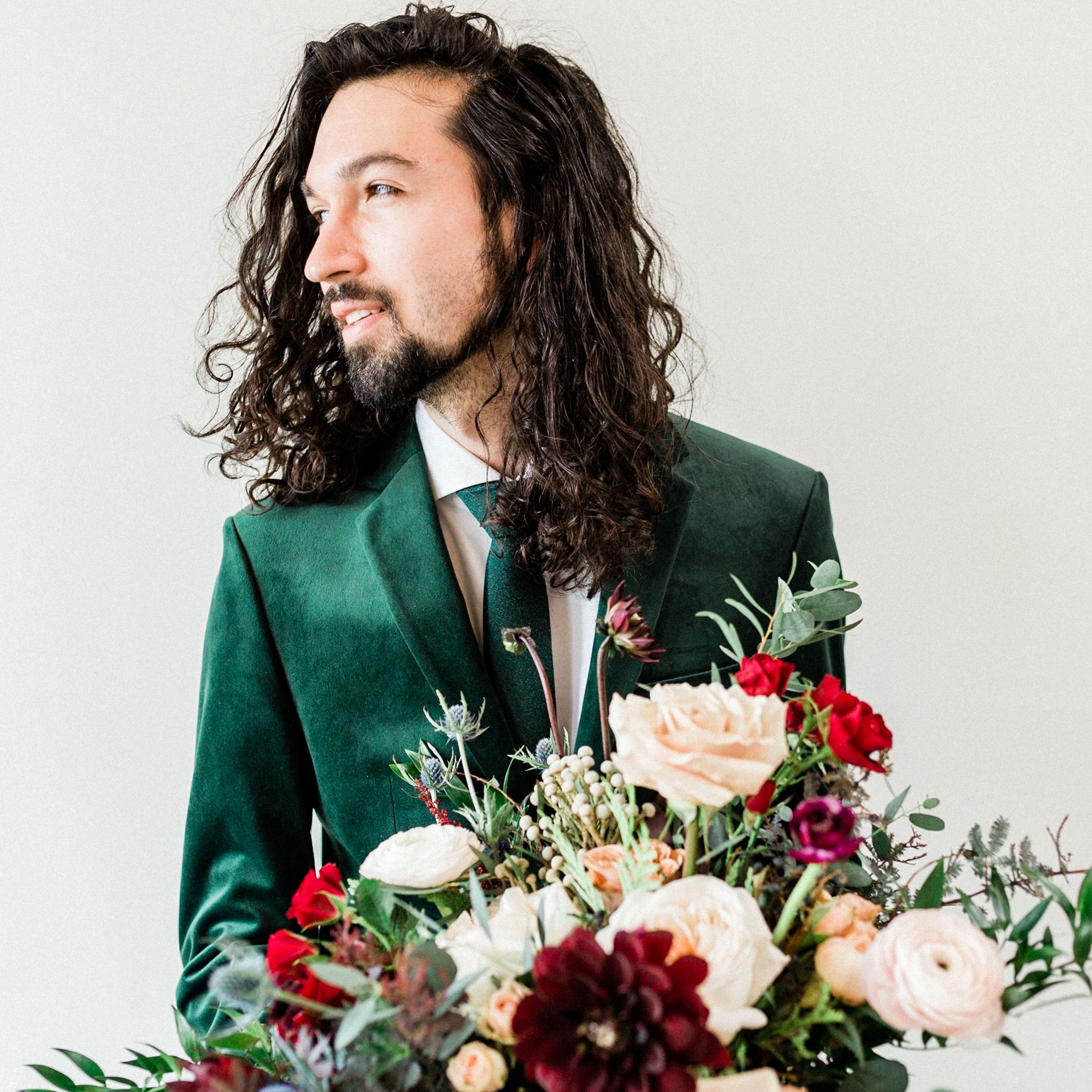 Image of Matthew Ramirez standing behind a bouquet of beautiful flowers wearing a green suit and tie. A white man with long dark brown hair with a beard and moustache looks off to the left into the distance.