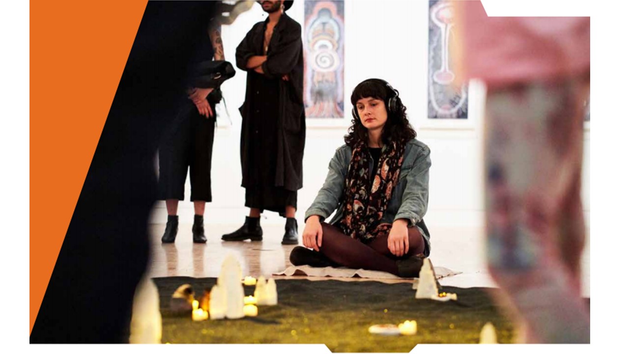 A woman sits crosselegged on a towel on the floor wearing headphones in front of a set of candles as other people mill around to the right hand side of the image chatting.