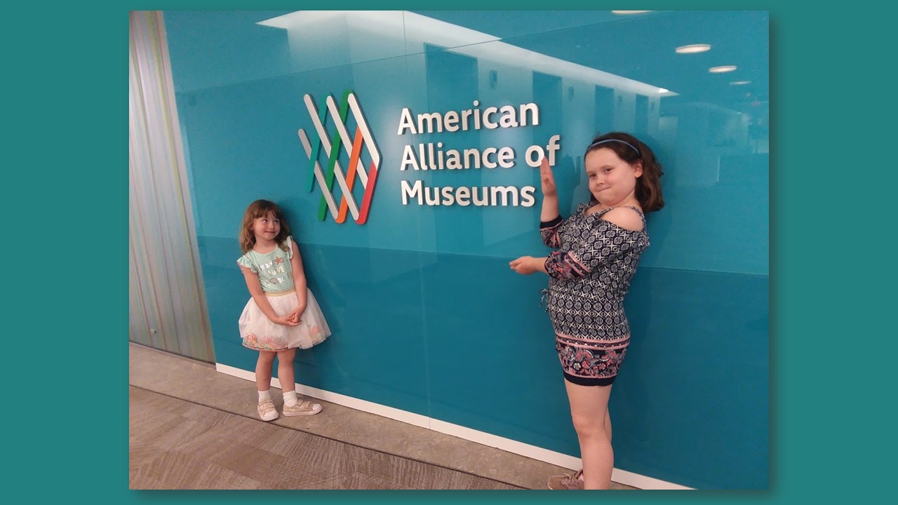 Two young white girls stand in front of the American Alliance of Museums logo and sign