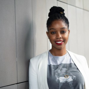 Headshot of Ravon Ruffin, a black woman with dark brown hair pulled into a topknot leaning against a stark concrete wall wearing a graphic t-shirt with a white jacket.
