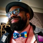 Image of Saleem Penny, a black man wearing a bowler hat, blue rimmed glasses and a color bow tie.