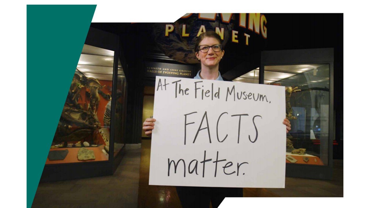 Emily Graslie, Field Museum Chief Curiosity Correspondent stands in front of an exhibtion on the planet earth with a handwritten sign that says 'At The Field Museum FACTS matter".