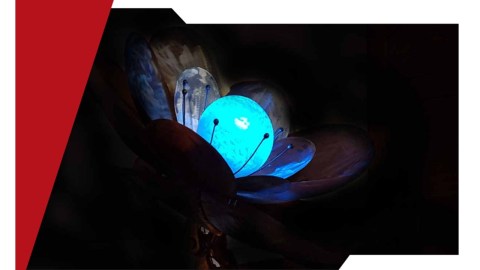 An electronic flower with metal petals sits in a dark room with a green glowing orb in the middle.