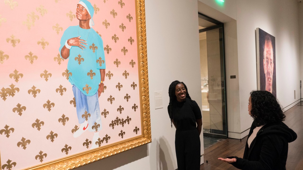 Asmaa stands in front of a bright blue and pink painting by Kehinde Wiley, engaging another person in conversation. She is a young black woman with braided hair and wears a black shirt and pants.