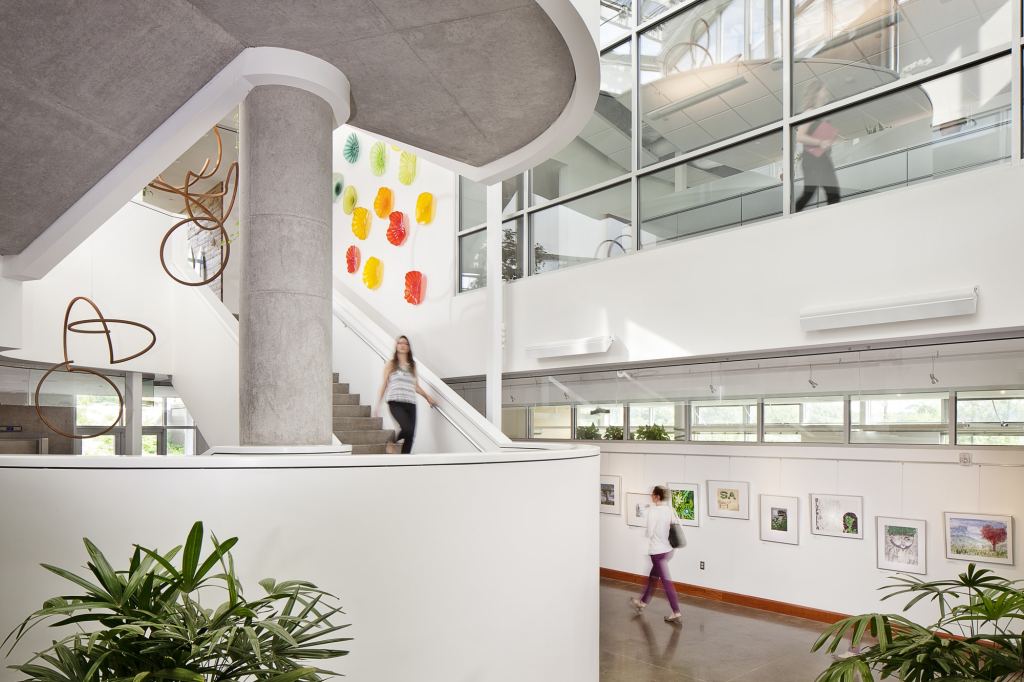 The lobby of a building with strong natural lighting, large glass windows, colorful art with organic shapes displayed, and plants on view.