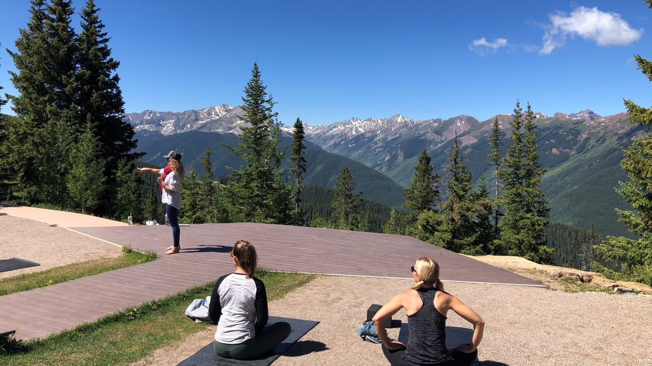 Three people are set up with yoga mats in the mountains, looking out over the landscape.