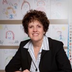 Image of Carole Charnow, a white woman sitting in front of a wall with children's drawings wearing a dark suit jacket and striped shirt. 
