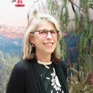 A white woman with shoulder length blond hair and red rimmed glasses stands in front of a natural backdrop.