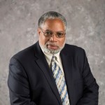 Image of Lonnie Bunch, a black man with silver and black hair, sits in front of a grey backdrop wearing a dark grey suit with a striped tie and metal framed glasses. 