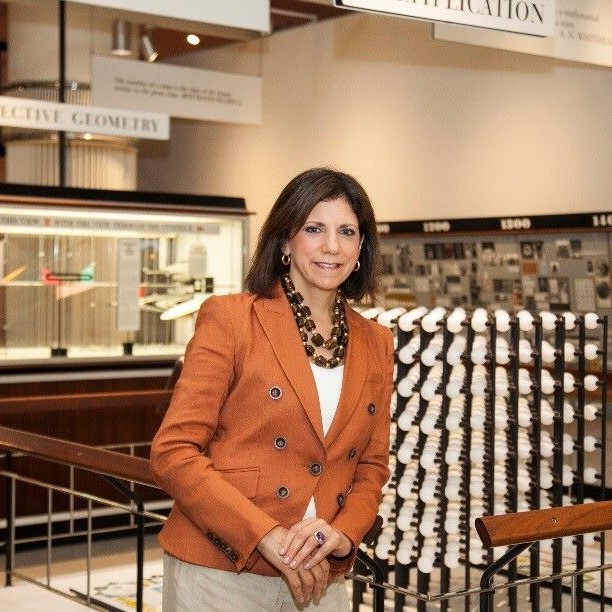 Image of a white woman, Patricia Mooradian, standing in a gallery wearing a peach colored jacket with dark brown hair down to her shoulders.