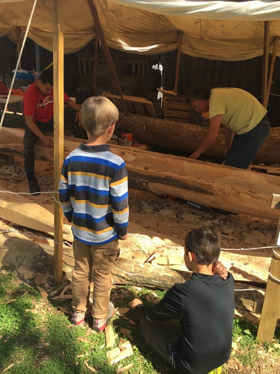 Young visitors watch two workers work to carve a large tree under a canopy tent outside. 