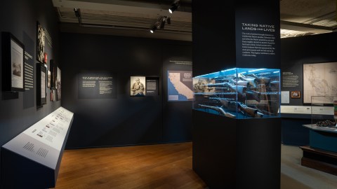 Interior view of the exhibition, with a display case of guns and the title "Taking Native Lands and Lives" visible