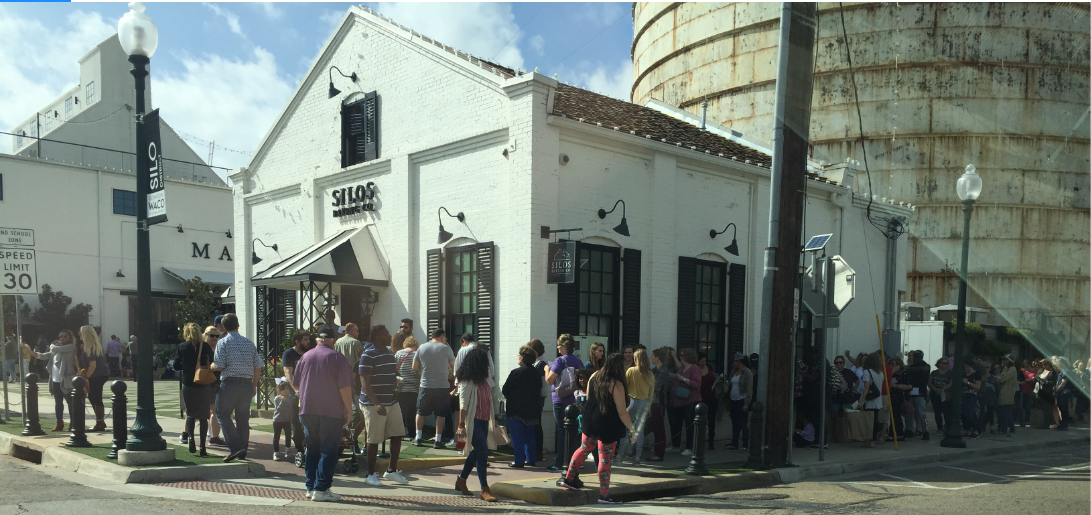 An exterior view of a building labeled "The Silos" in front of a grain silo, with a crowd of people lined up outside.