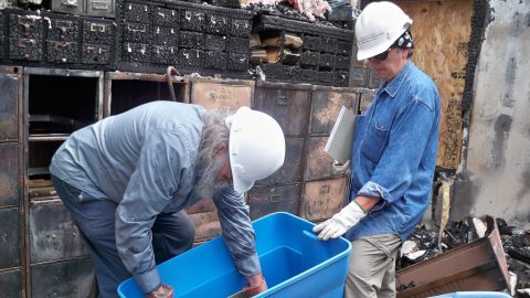 Two people stand in the midst of a destroyed collections facility, wearing hard hats and carrying a plastic bin for retrieving salvageable items.