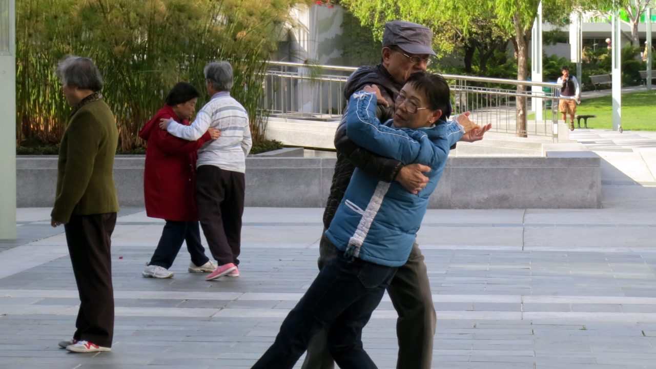 Pairs of older adults dance in an outdoor park.
