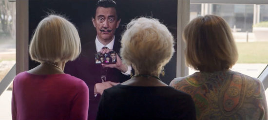 A group of three visitors is seen looking at the AI Dali, who holds a smartphone displaying a group selfie of himself and the visitors.