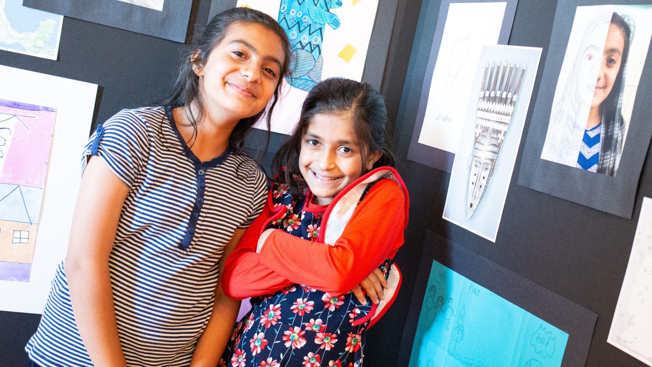 Two children pose in front of a gallery of art projects, including self-portraits and illustrations.