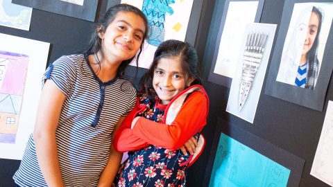 Two children pose in front of a gallery of art projects, including self-portraits and illustrations.