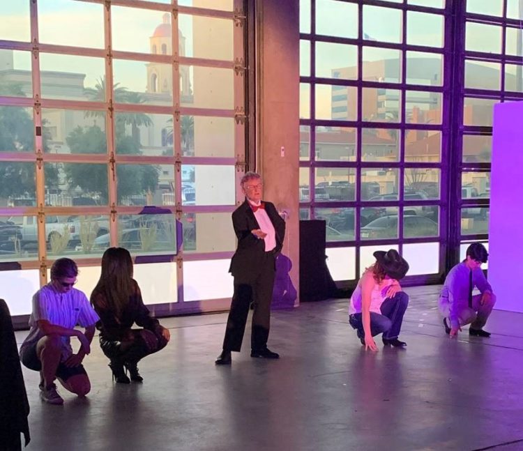 A drag king, in a suit and bowtie, stands center stage surrounded by other kneeling performers.