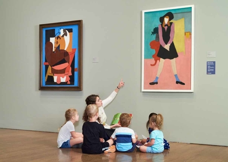 A group of children is seen in front of the painting the label describes with an adult guide. The painting is of a young woman, rendered in flat blocks of color and stylized forms.