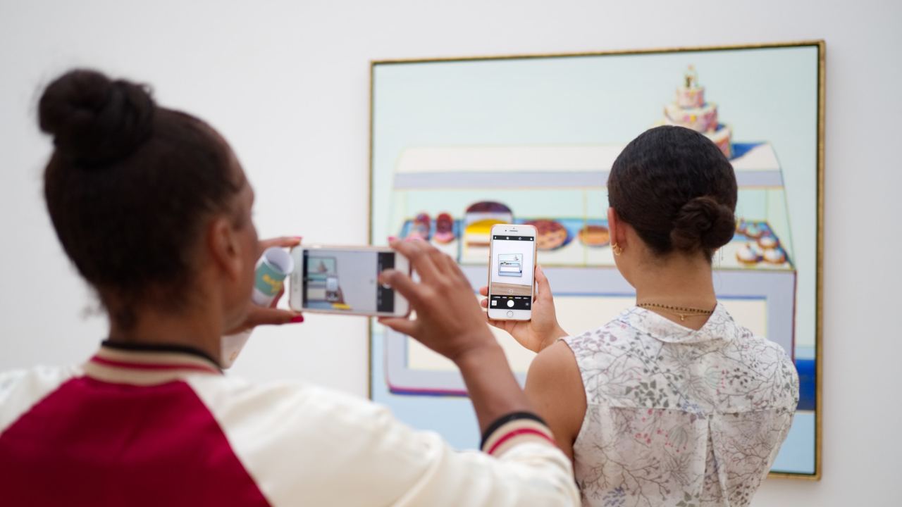 Two museum visitors hold take pictures of a Wayne Thiebaud painting on their smartphones.
