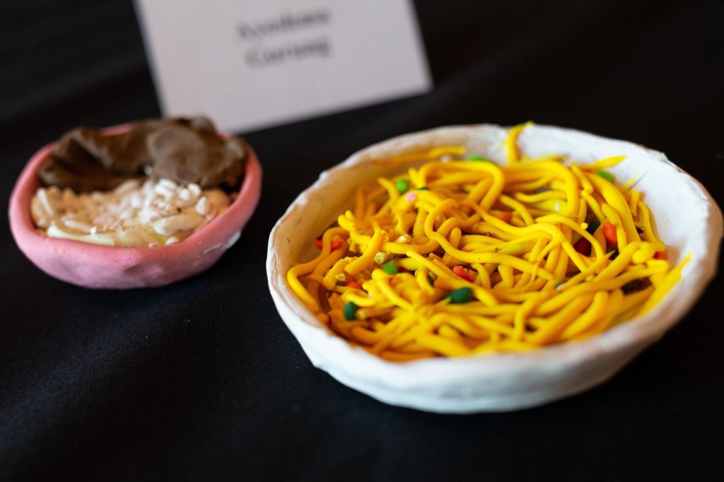 Two clay sculptures of bowls of food, one bright yellow noodles and the other a mixture of leaves and roots.