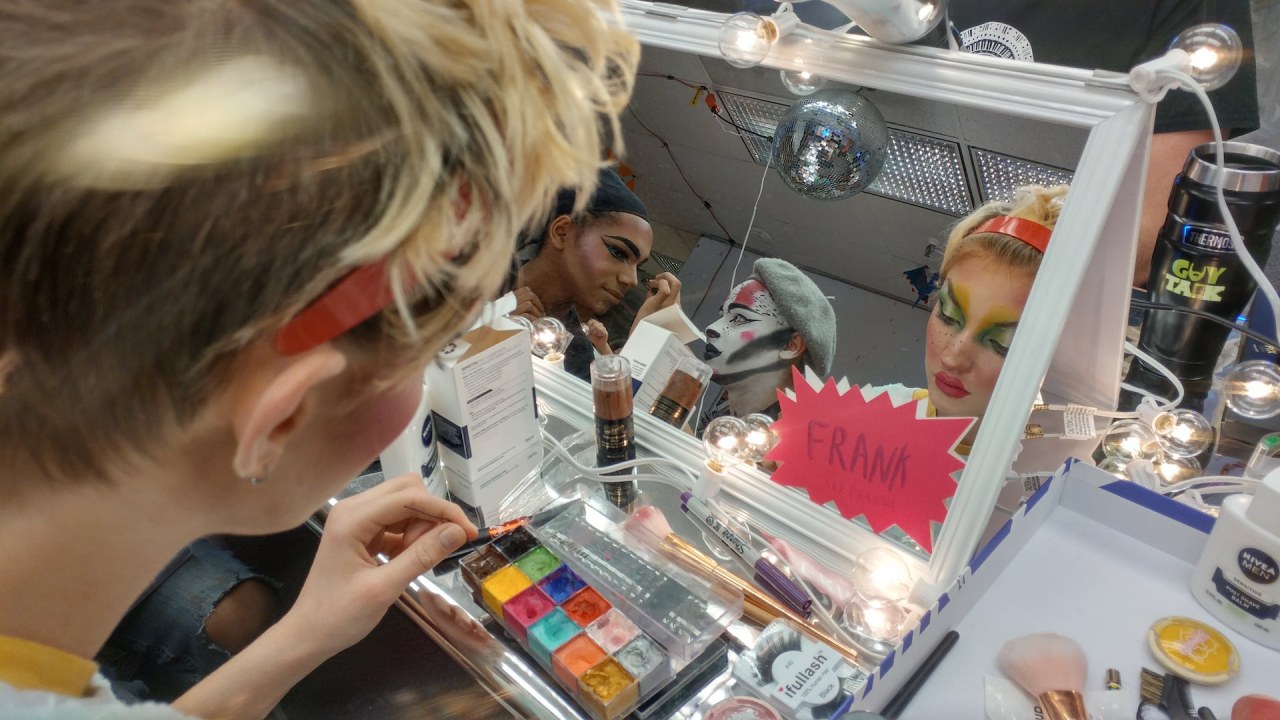 The performers are seen in a vanity mirror applying colorful and extravagant makeup looks.