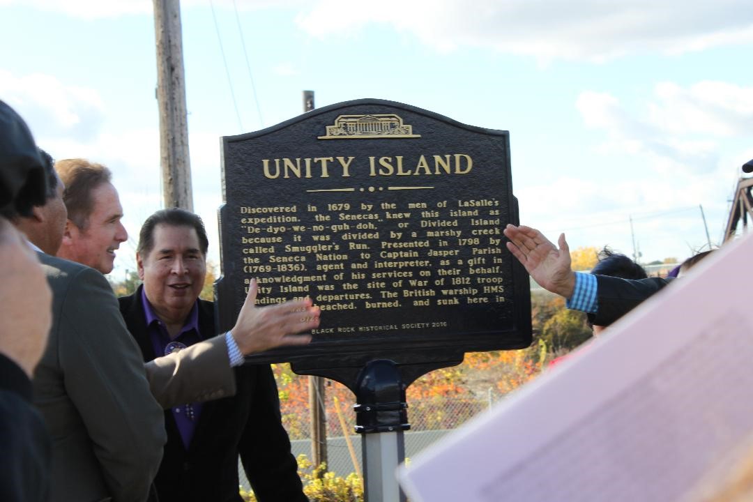 A group of people gather around a historical marker with the name "Unity Island" engraved on it.