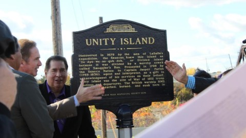 A group of people gather around a historical marker with the name "Unity Island" engraved on it.
