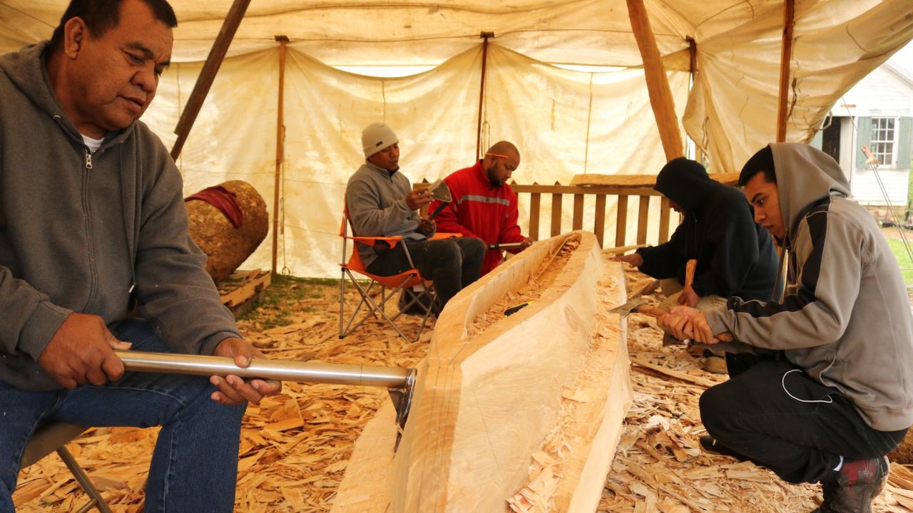 Liton and a group of four other people are seen under a tent carving the canoe, surrounded by a pile of wood shavings.