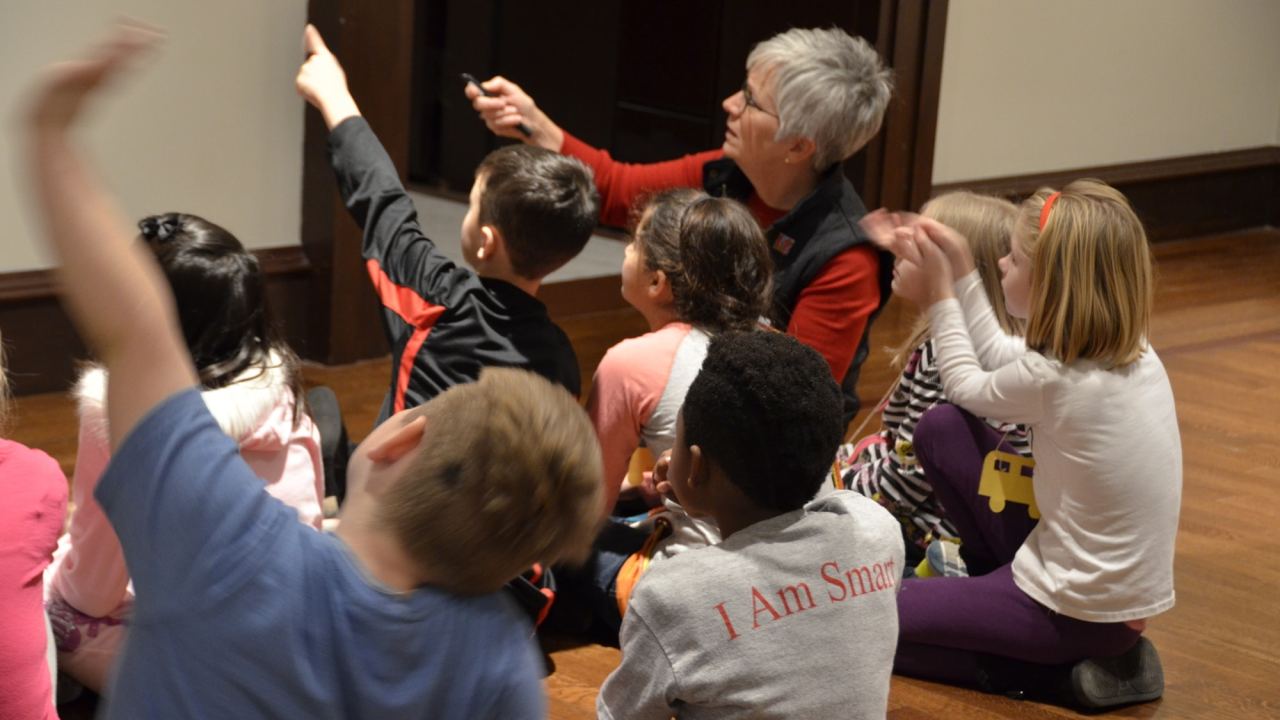 A docent is seen with a group of children seated on the floor, some of whom point in front of them to an artwork.