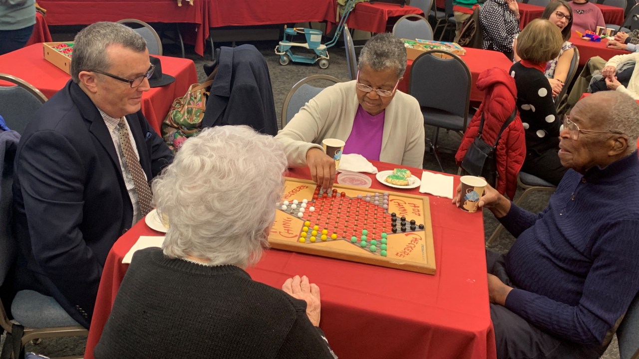 Four people sit around a table playing "Chinese checkers." Other tables of people are seen in the background.