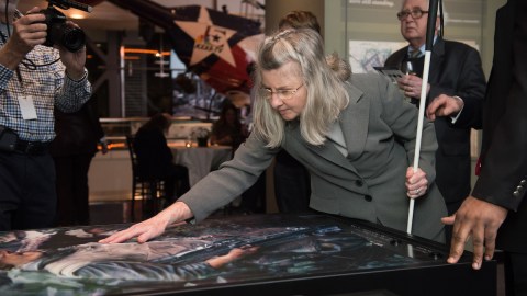 A woman holding a cane leans over a flat table with her fingertips grazing the raised surface of a print.